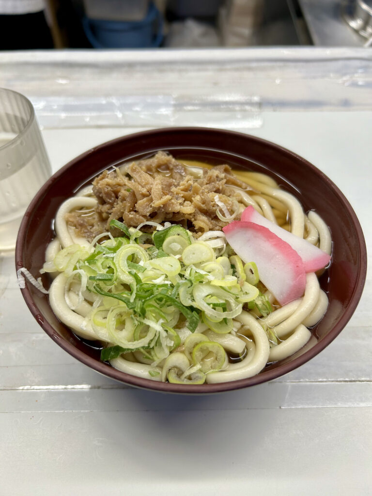 小倉駅のホームで提供される名物「かしわうどん」。茶色の丼に盛られた柔らかなうどんの上に、甘辛く煮込まれた鶏肉（かしわ）、刻みネギ、紅白のかまぼこがトッピングされている。九州らしい甘めの出汁が並々と注がれており、横にはお冷のコップが置かれている。