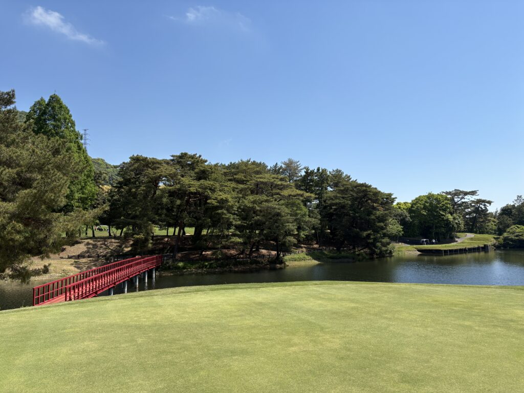 門司ゴルフ倶楽部18番ホールのグリーン上からティーグラウンド方向を振り返った景色。手前には美しく刈り込まれたグリーンの芝生が広がり、左手には池に架かる鮮やかな赤い橋が見える。池の対岸には深い緑の木々が茂り、その奥にティーグラウンドが位置している。青空の下、静寂と格式を感じさせる名門コースの風景。
