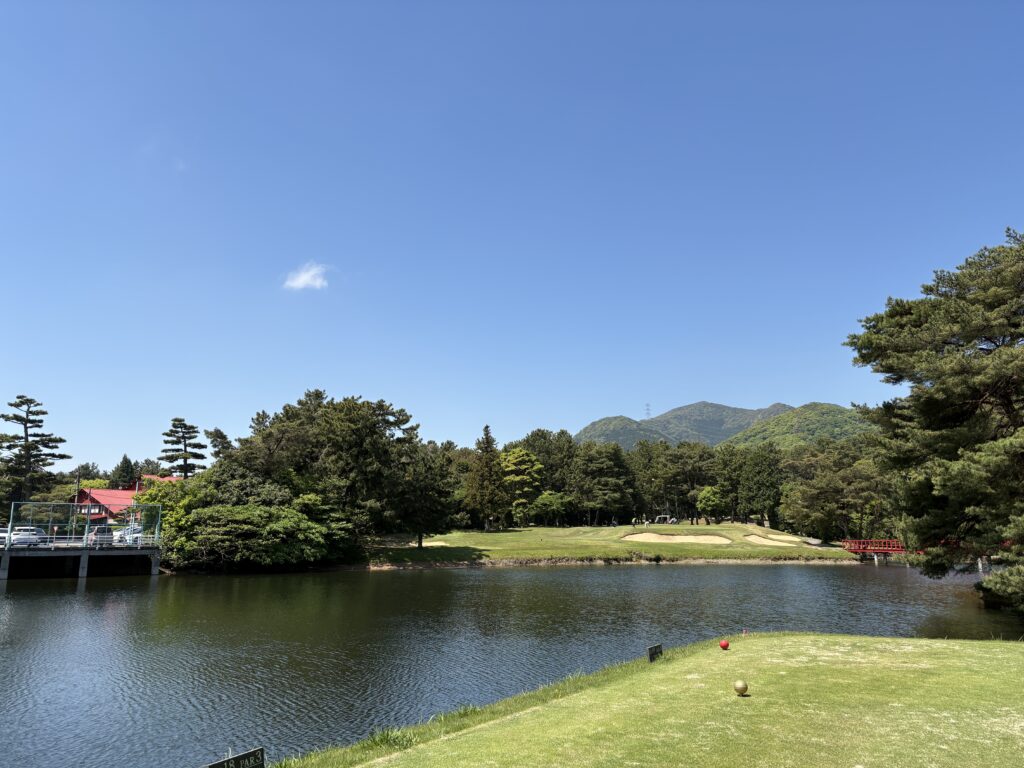門司ゴルフ倶楽部18番ホール、池越えのショートホールのティーグラウンドからの眺め。手前の芝生にはティーマークが置かれ、目の前には大きな池が広がる。池の向こう、左側には特徴的な赤い屋根のクラブハウス、右側には赤い橋が緑の木々に映えている。奥には山並みと青空が広がり、池の青、芝の緑、建物と橋の赤のコントラストが美しい。